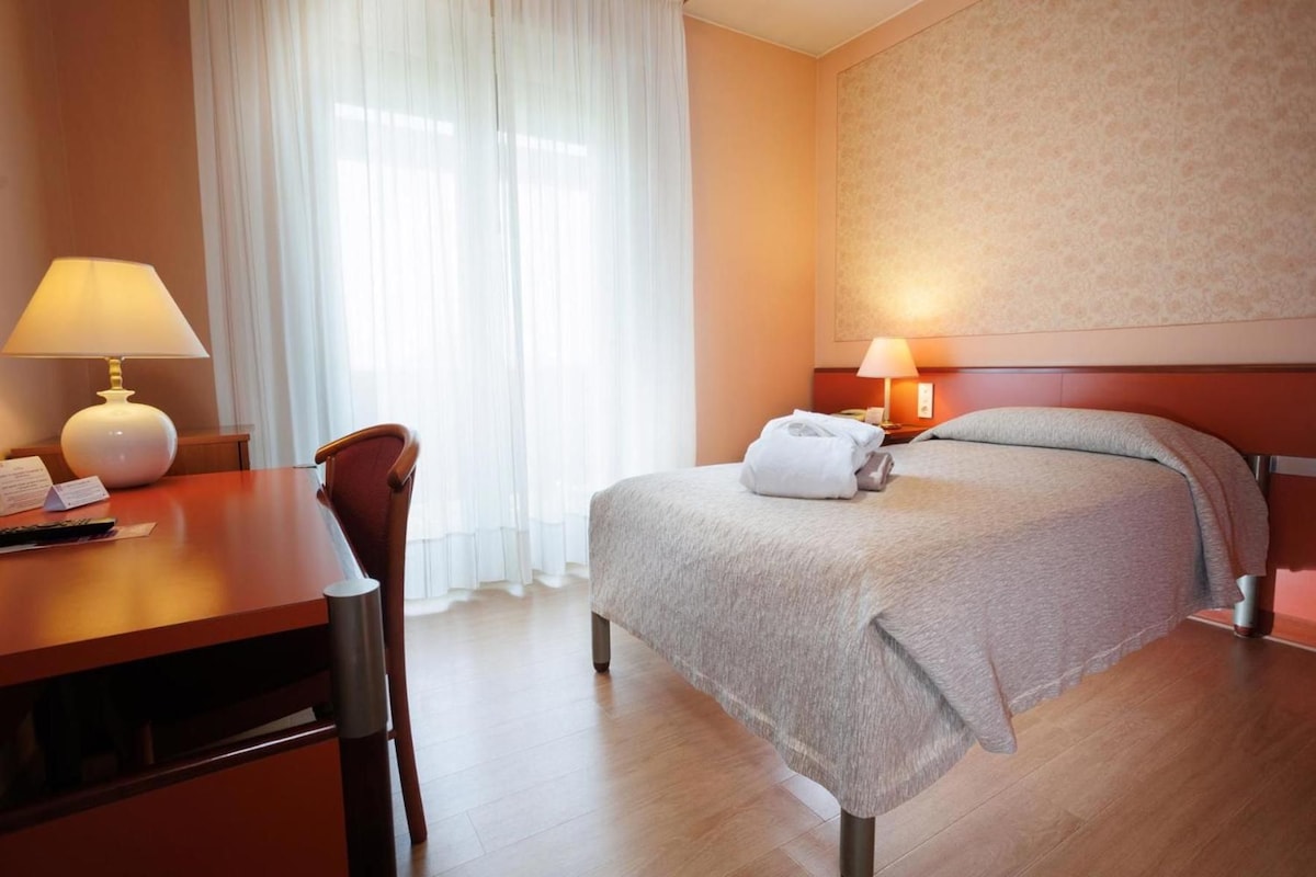 A hotel room with a bed, a desk, a chair, a lamp, and a window with curtains.