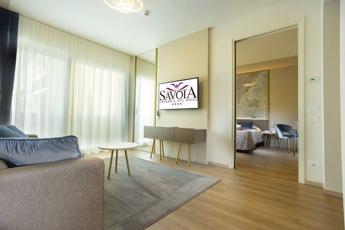 Une chambre d’hôtel moderne, équipée d’un canapé, d’une petite table ronde, d’un lit avec une tête de lit et d’un téléviseur fixé au mur, sur lequel est affiché le nom de l’hôtel.