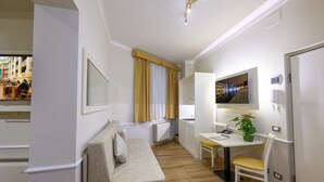 Moderni hotellihuone, jossa on sohva, ruokapöytä ja tuoli, pieni työp öytä, ruukkukasvi ja suuri ikkuna, jossa on verhot.