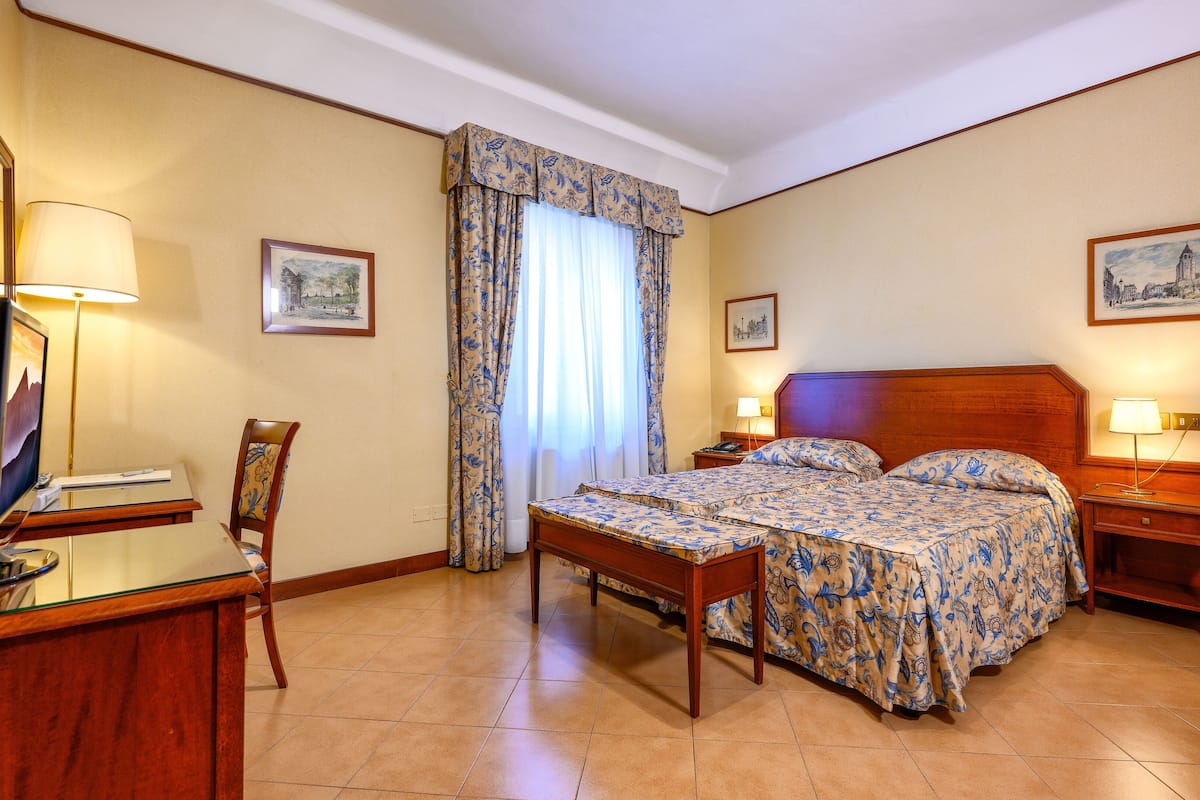 Ein Hotelzimmer mit einem Bett, Nachttischen, einem Schreibtisch mit Computer, einem Sessel und zwei Bildern an der Wand.