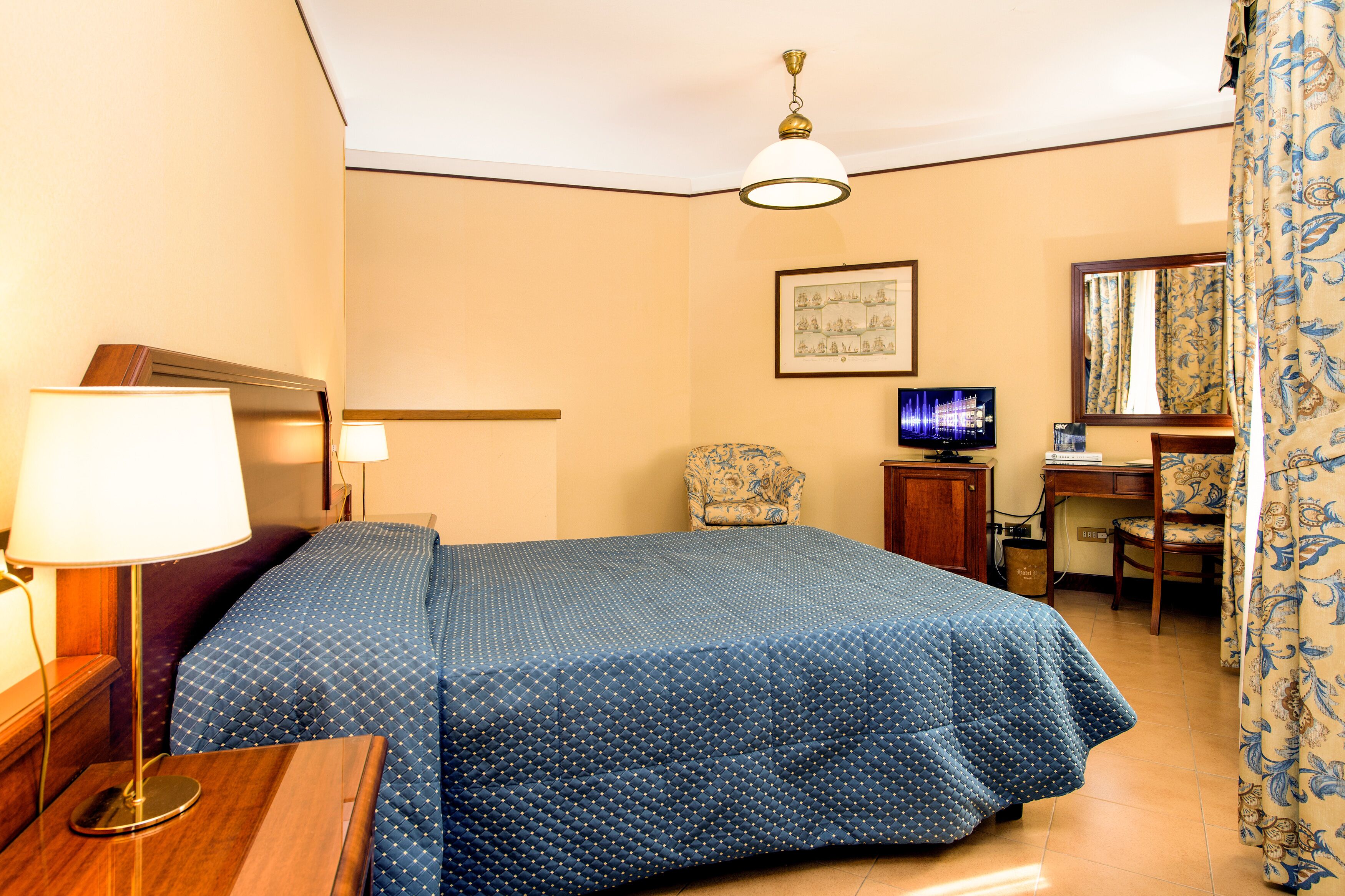 A hotel room with a bed, a desk, a chair, a lamp, and a television.