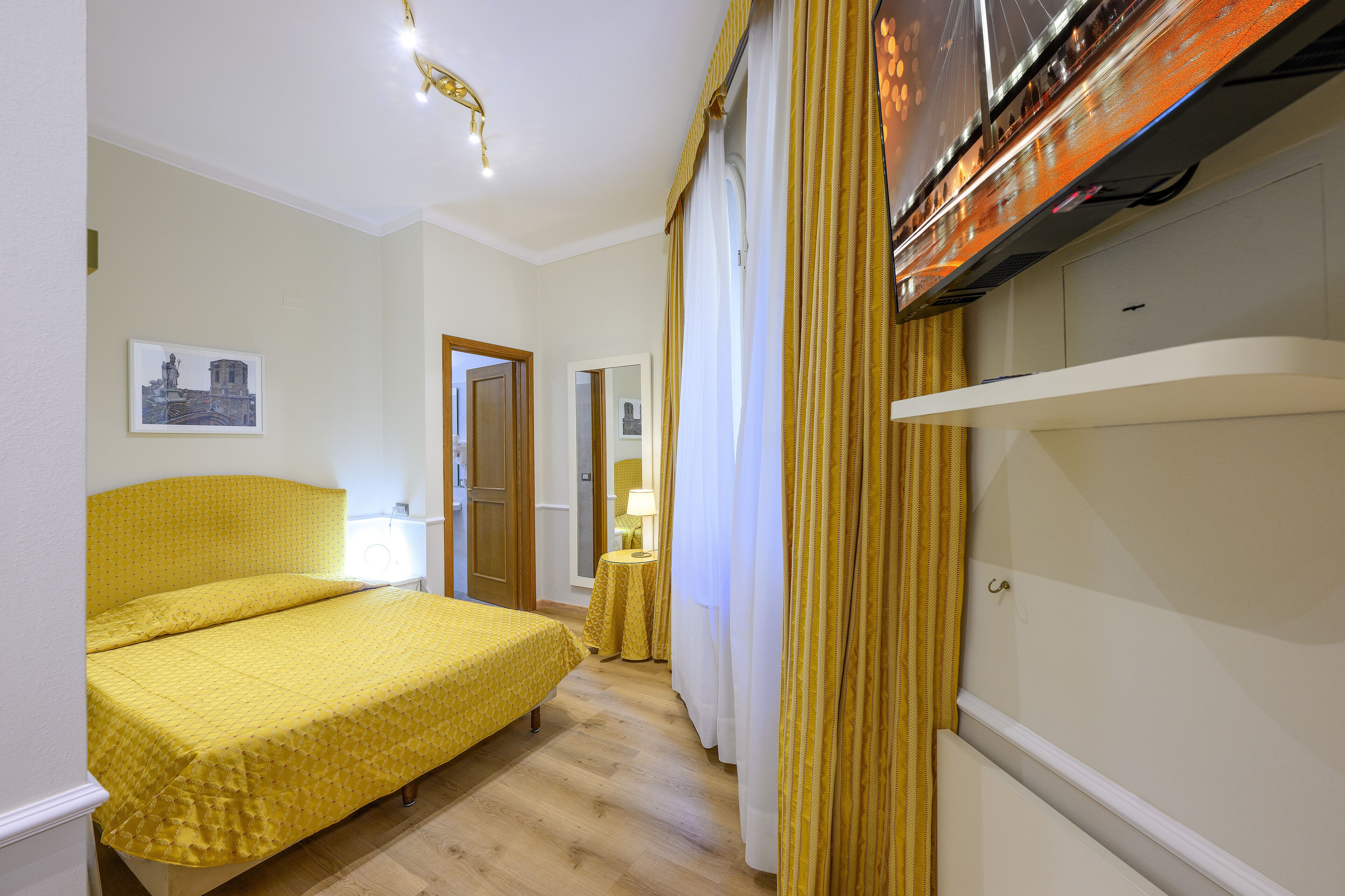 Una camera d'albergo con un letto, un tavolino con una lampada e una televisione appesa al muro.
