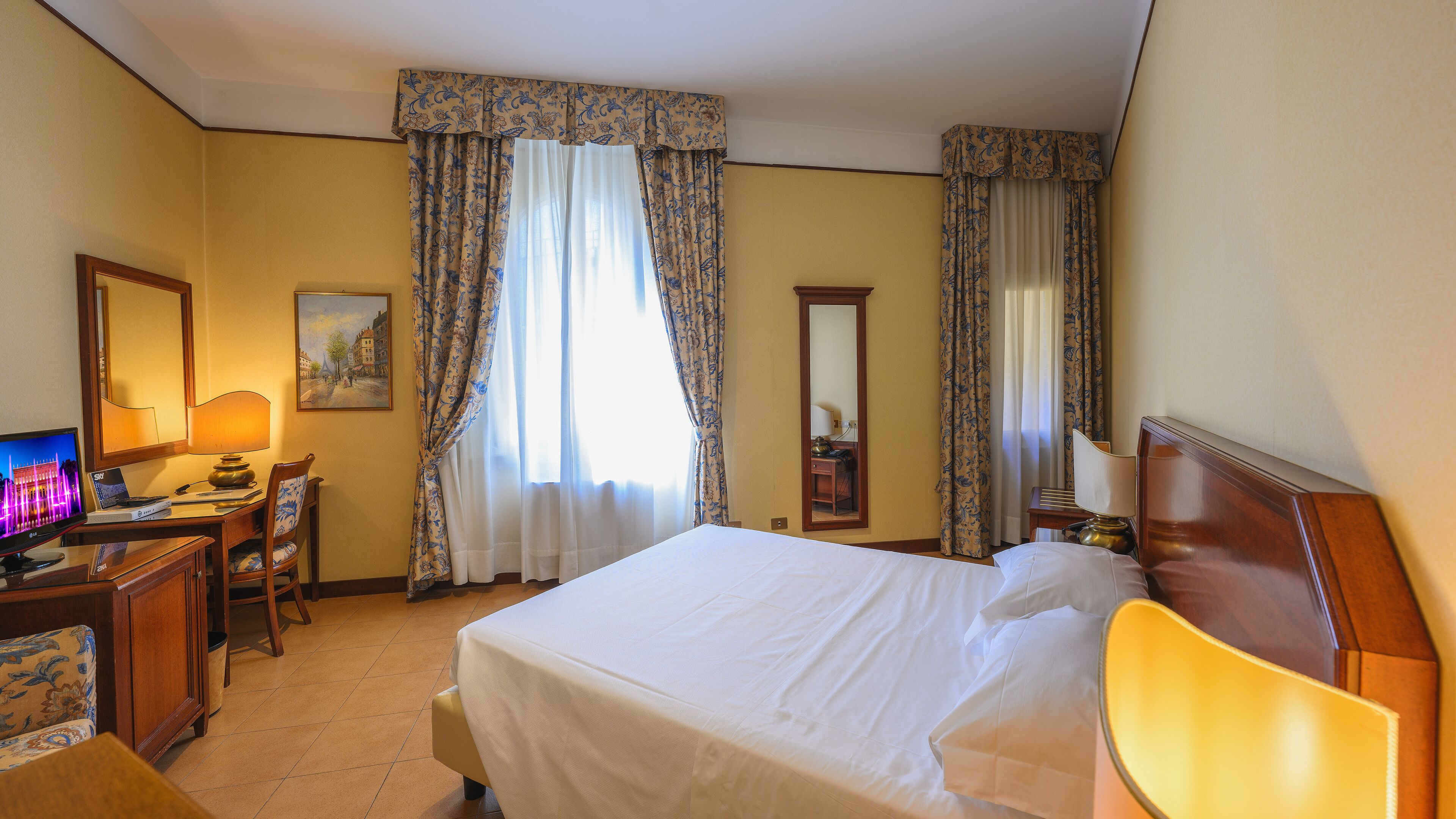 Une chambre d’hôtel avec un lit, un bureau doté d’une lampe, une chaise, un téléviseur et des rideaux.