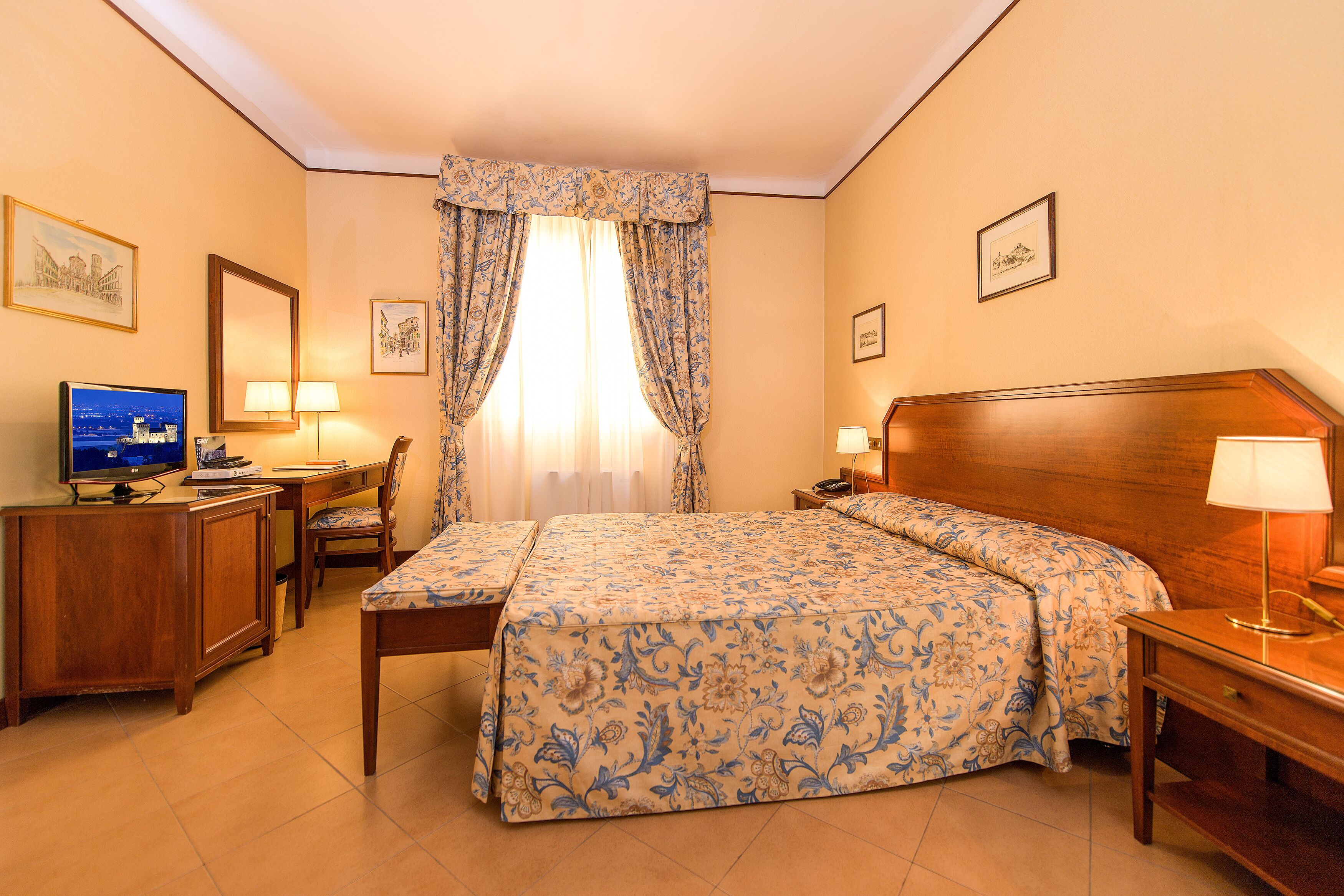 Quarto de hotel com cama, escrivaninha, cadeira e televisão.