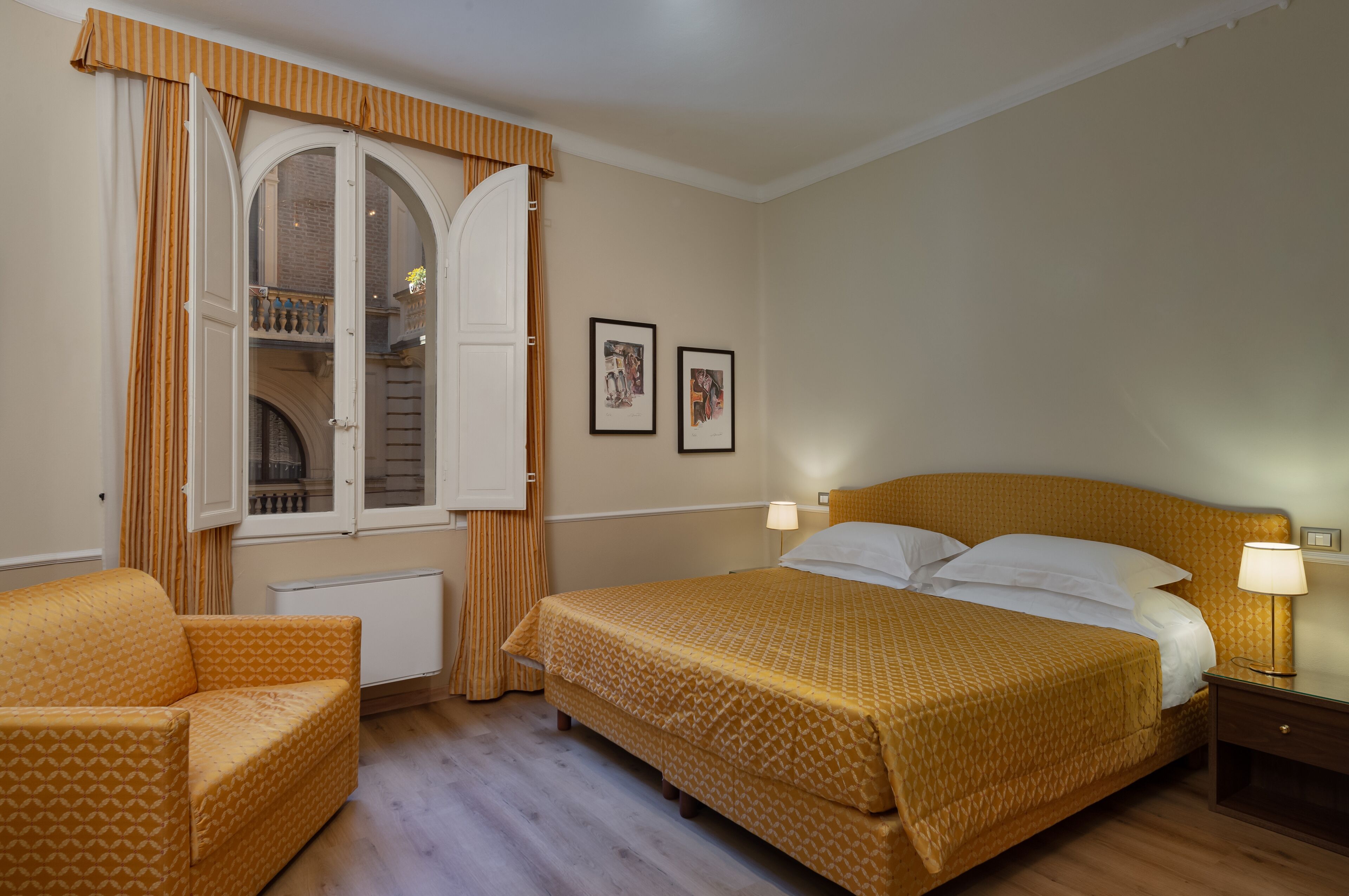 A hotel room with a bed, a chair, a window with curtains, and two framed pictures on the wall.