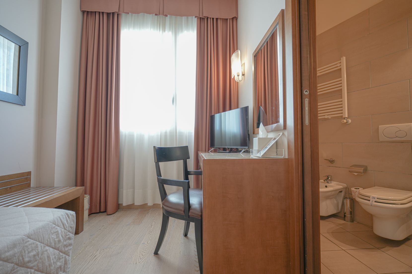 Habitación de hotel con cama, escritorio con computadora, silla, televisor, baño con inodoro y lavamanos, y ventana con cortinas.