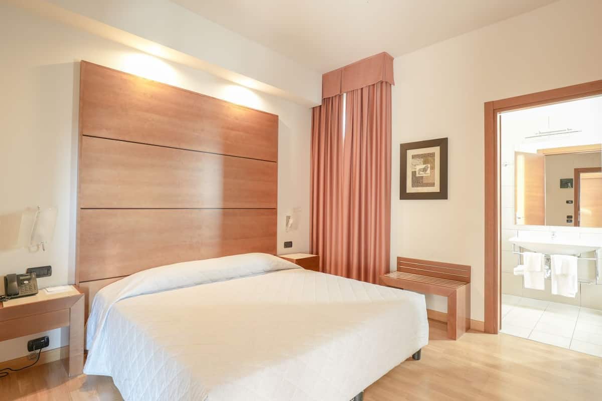 A hotel room with a large bed, a bedside table with a phone, a bench, a wall-mounted light, and a bathroom visible through an open door.