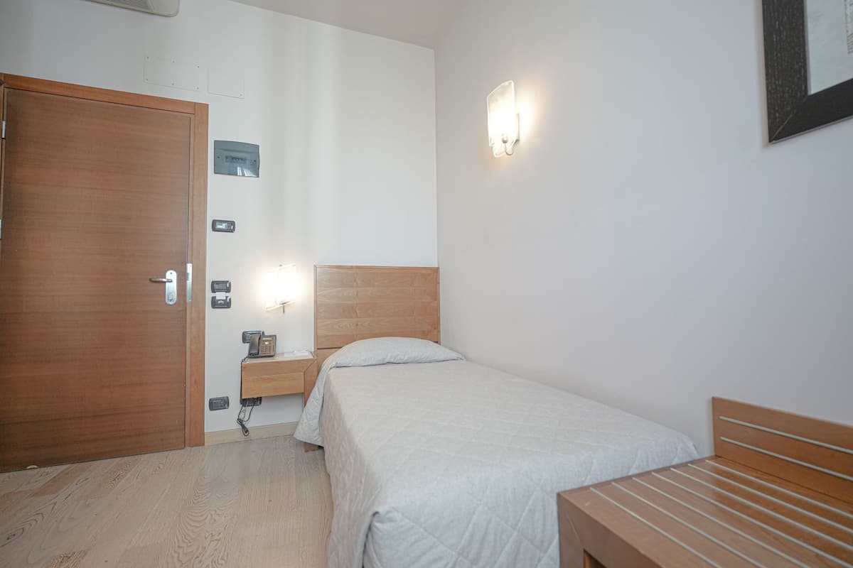 A small, clean room with a wooden door, a bedside table with a lamp, and a single bed.