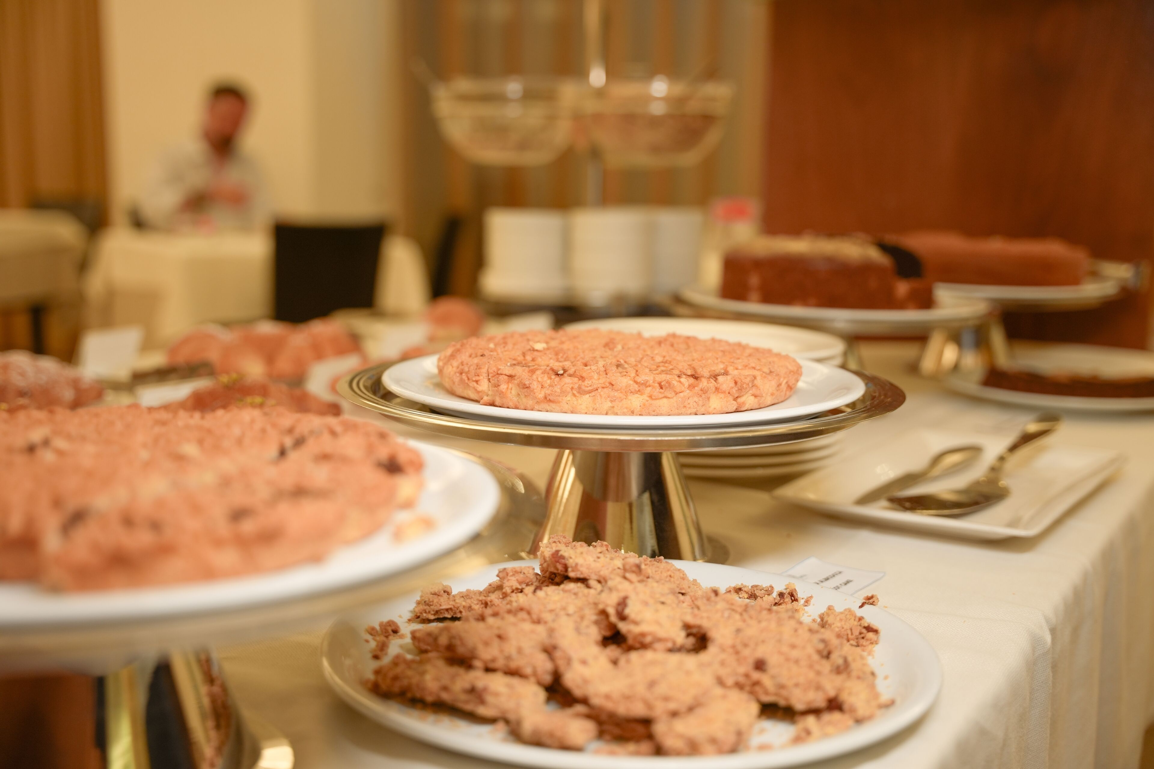 A buffet table with various dishes, including a large round cake on a golden stand, and a person in the background.