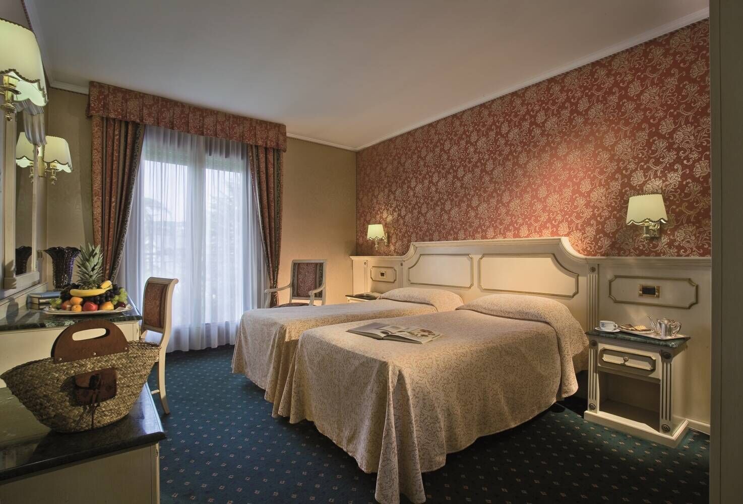 A hotel room with two beds, a desk with a fruit bowl, a chair, a dresser, and a patterned wallpaper.