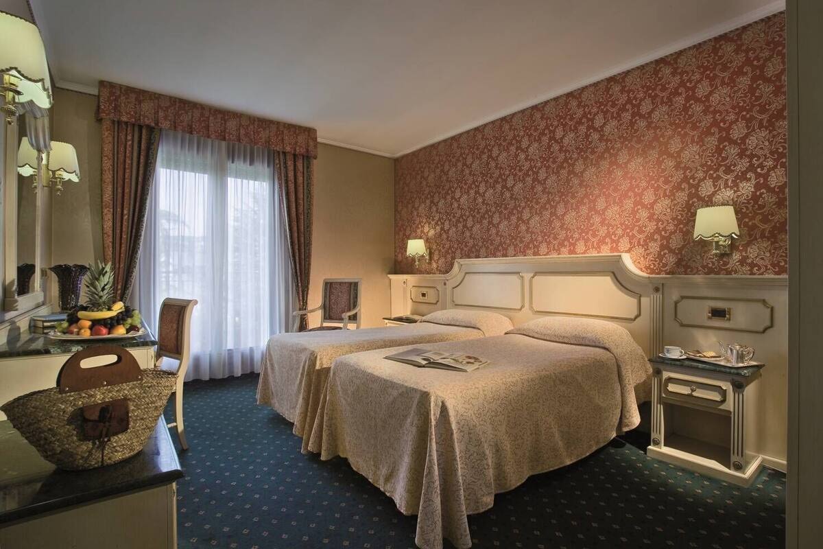 A hotel room with two beds, a desk with a fruit bowl, a chair, a dresser, and a patterned wallpaper.