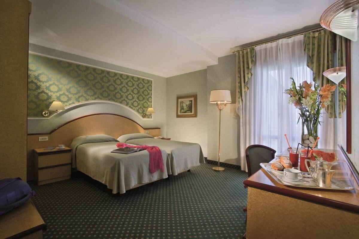 A hotel room with a bed, bedside tables, a desk with a tray of breakfast items, a lamp, and a window with curtains.