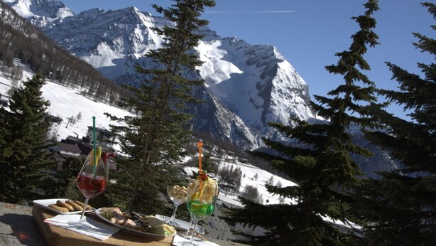 Ein Picknick mit Speisen und Getränken auf einem Tisch, mit Blick auf ein verschneites Gebirge.