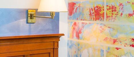 A wall-mounted lamp with a white shade, a wooden cabinet with a small drawer, and a colorful abstract painting.