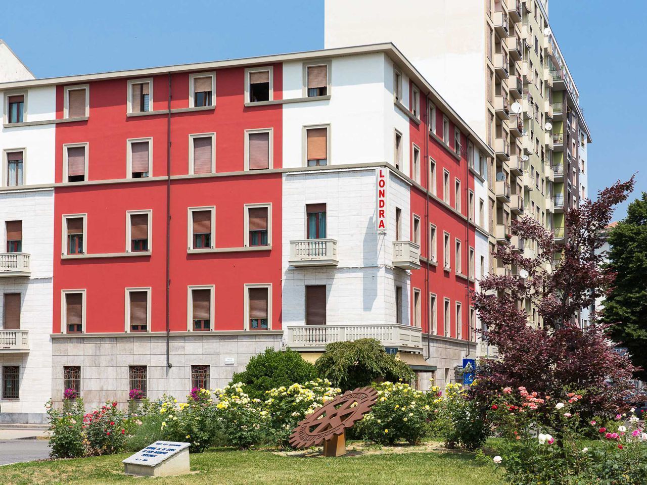 A multi-story building with a red and white facade, balconies, and a garden area with flowers.