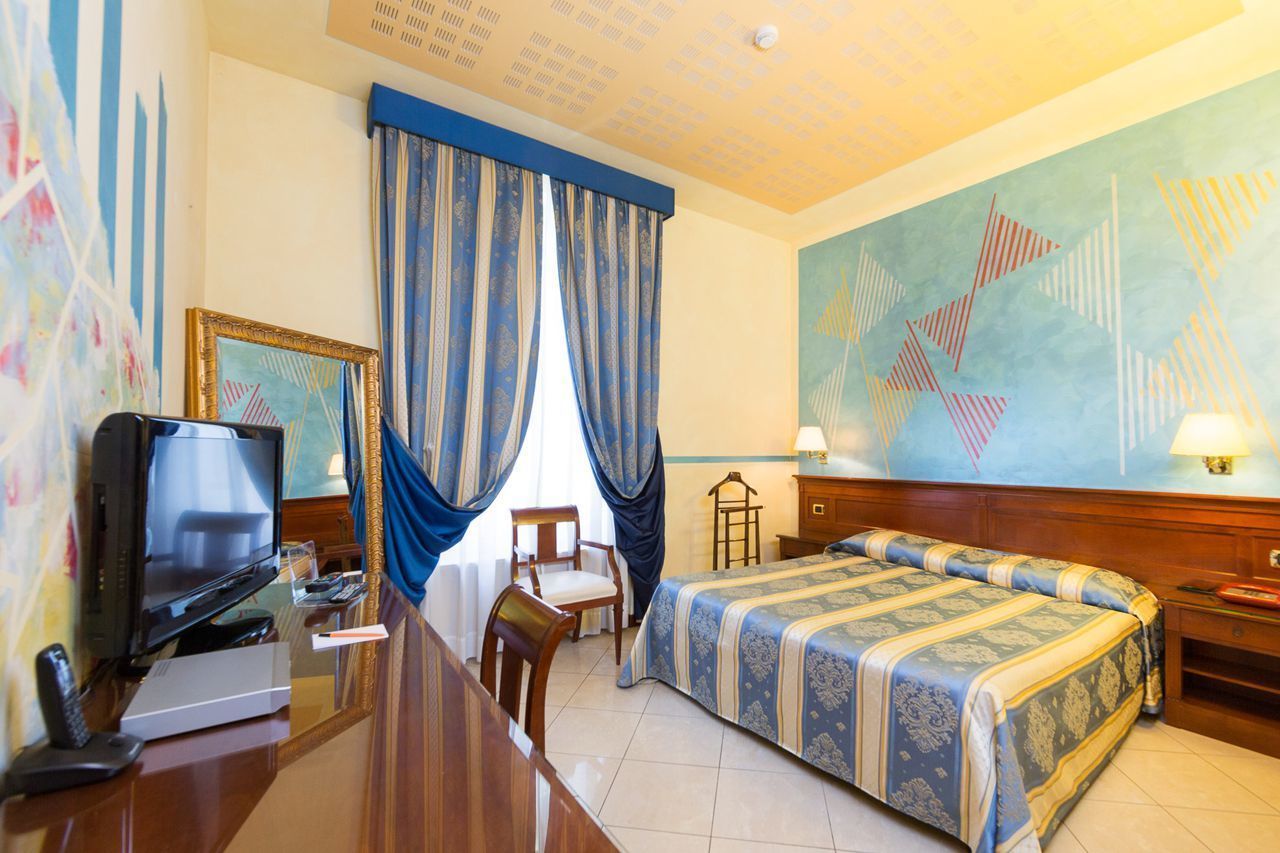 Habitación de hotel con cama, escritorio con televisor, una silla y un cuadro abstracto grande en la pared.