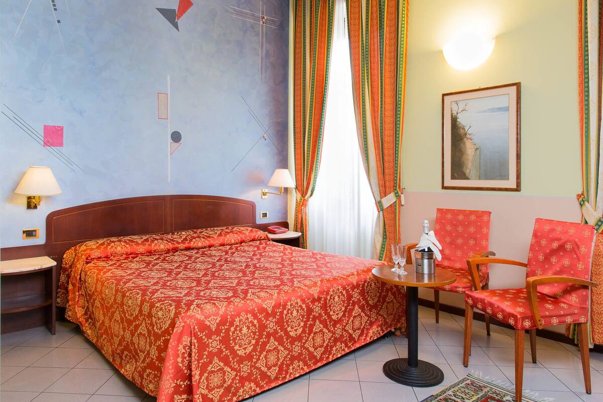 Ein Hotelzimmer mit einem großen Bett, einem kleinen Tisch mit Flasche und Gläsern, zwei Stühlen und einem Bild an der Wand.
