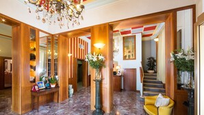 A lobby with a marble floor, wooden paneling, a yellow sofa, and a chandelier. - Hotel Londra (Alessandria)