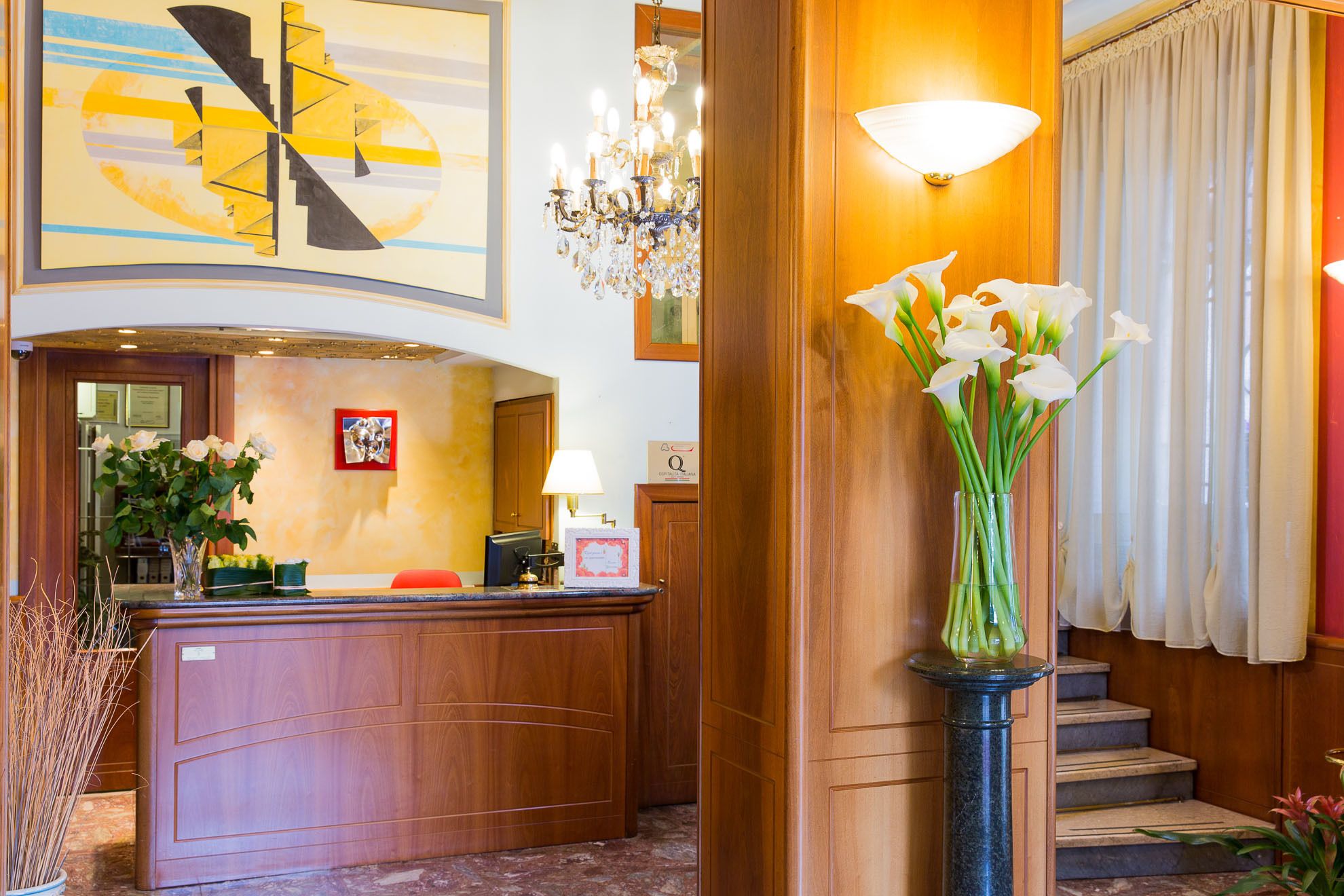 A hotel reception area with a wooden reception desk, a chandelier, and a vase of white flowers.