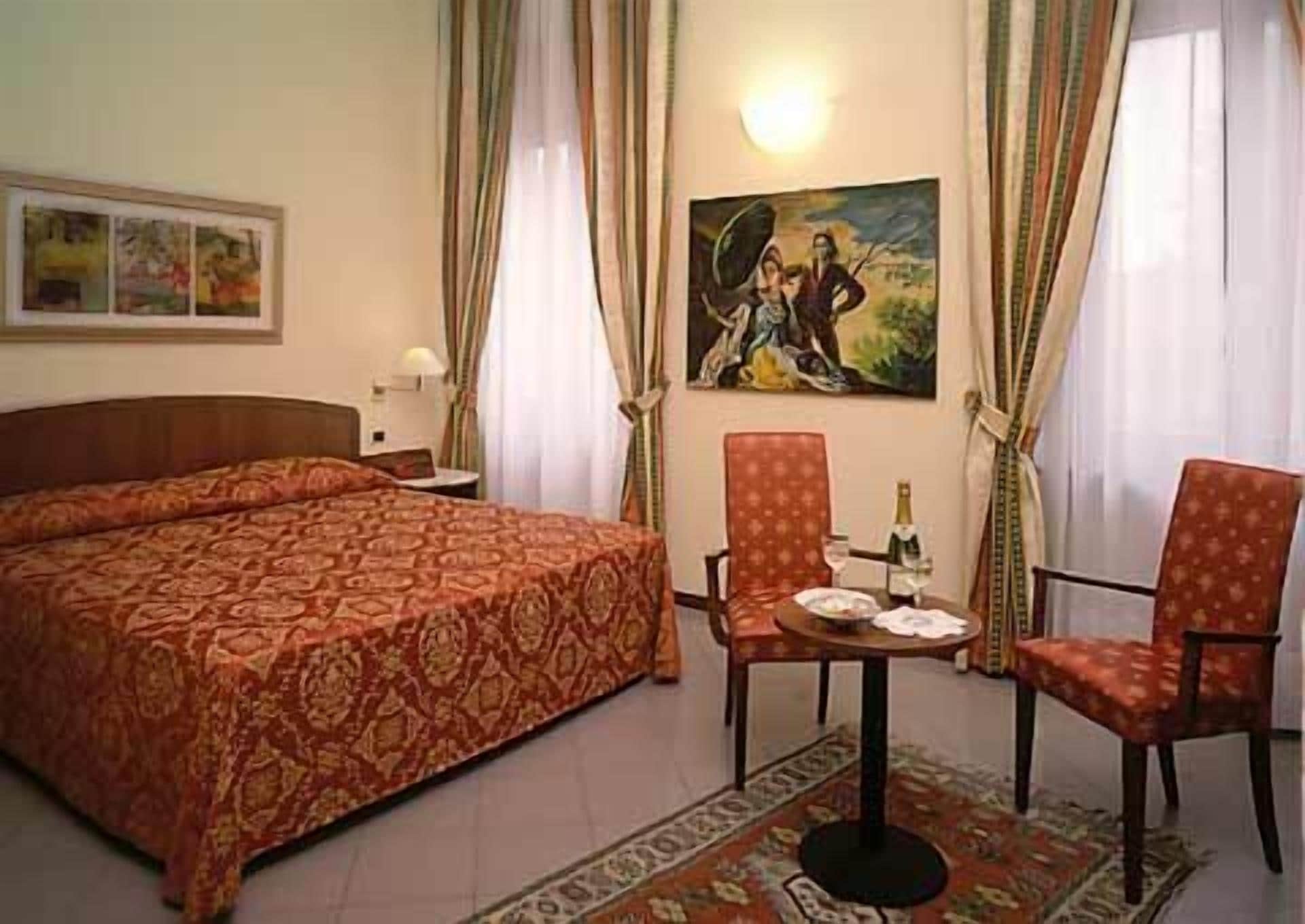 A hotel room with a large bed, two armchairs, a small table with a bottle and glasses, a painting on the wall, and curtains.