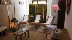 Una habitación con una mesa de madera, dos sillones de descanso, una mesita con una lámpara y un espejo decorativo.