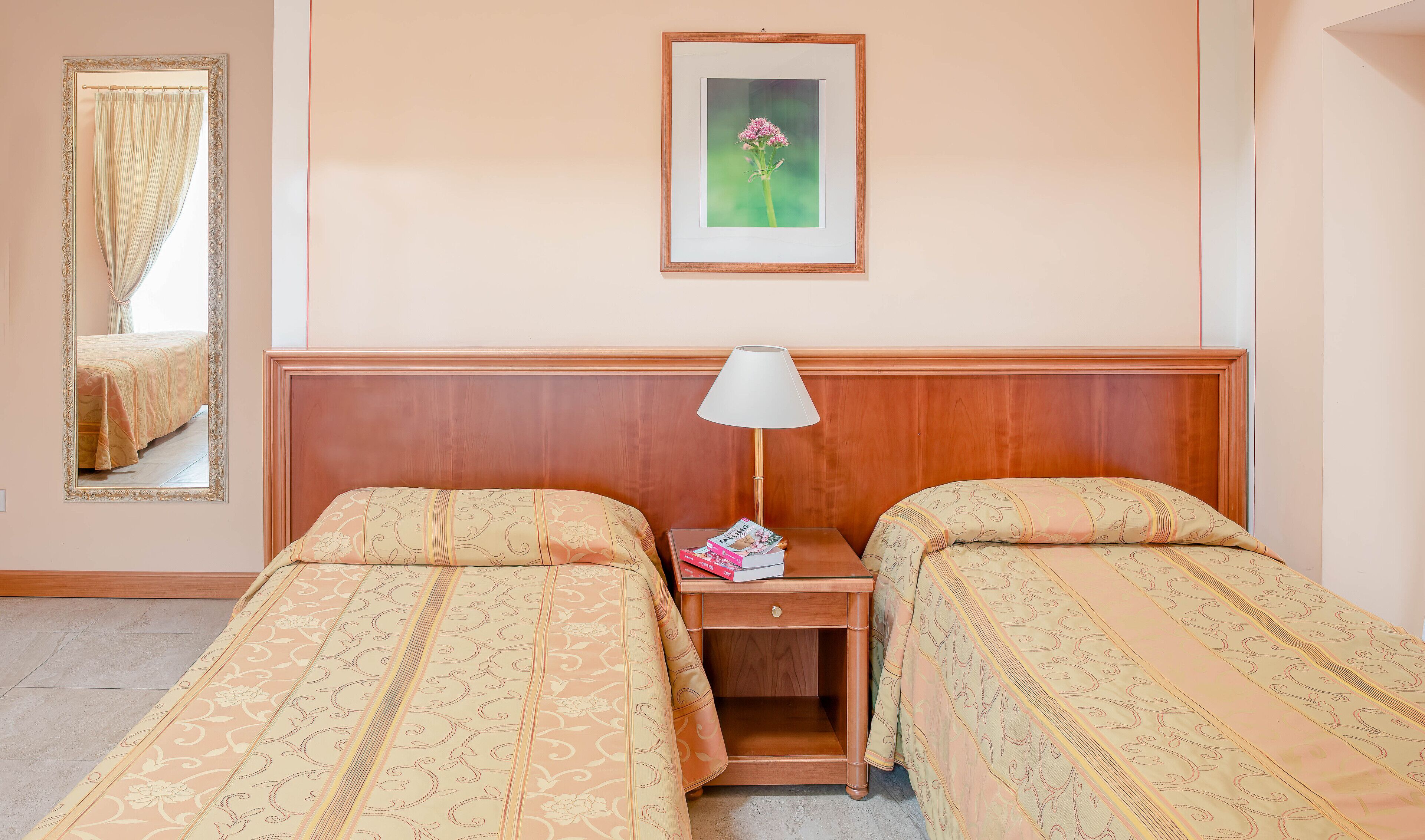 A hotel room with two single beds, a nightstand with a lamp, a framed picture on the wall, and a mirror on the left.