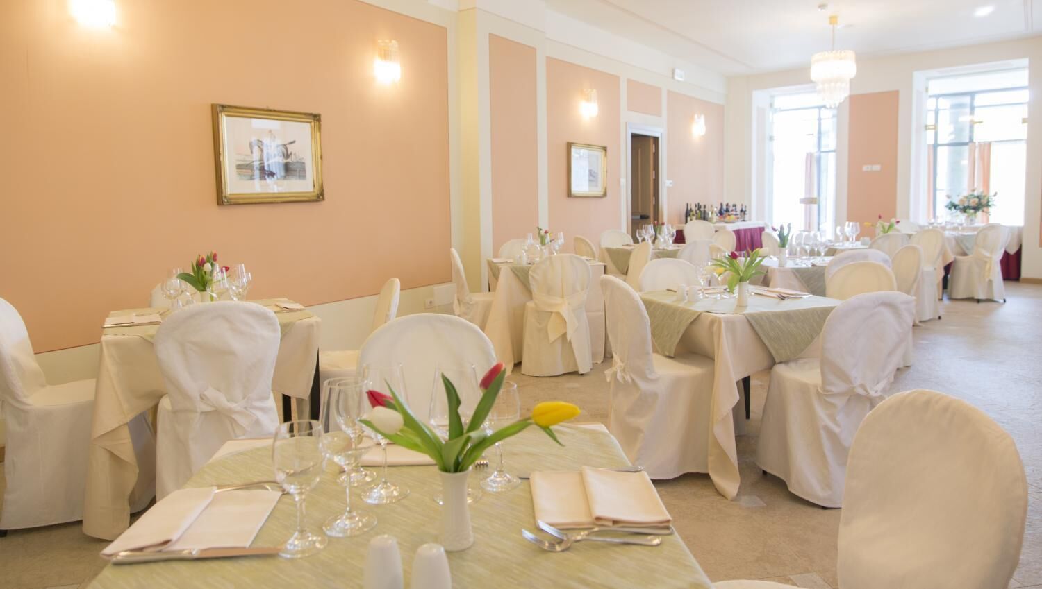 Un salón de banquetes con varias mesas dispuestas para comer, sillas blancas y una araña.