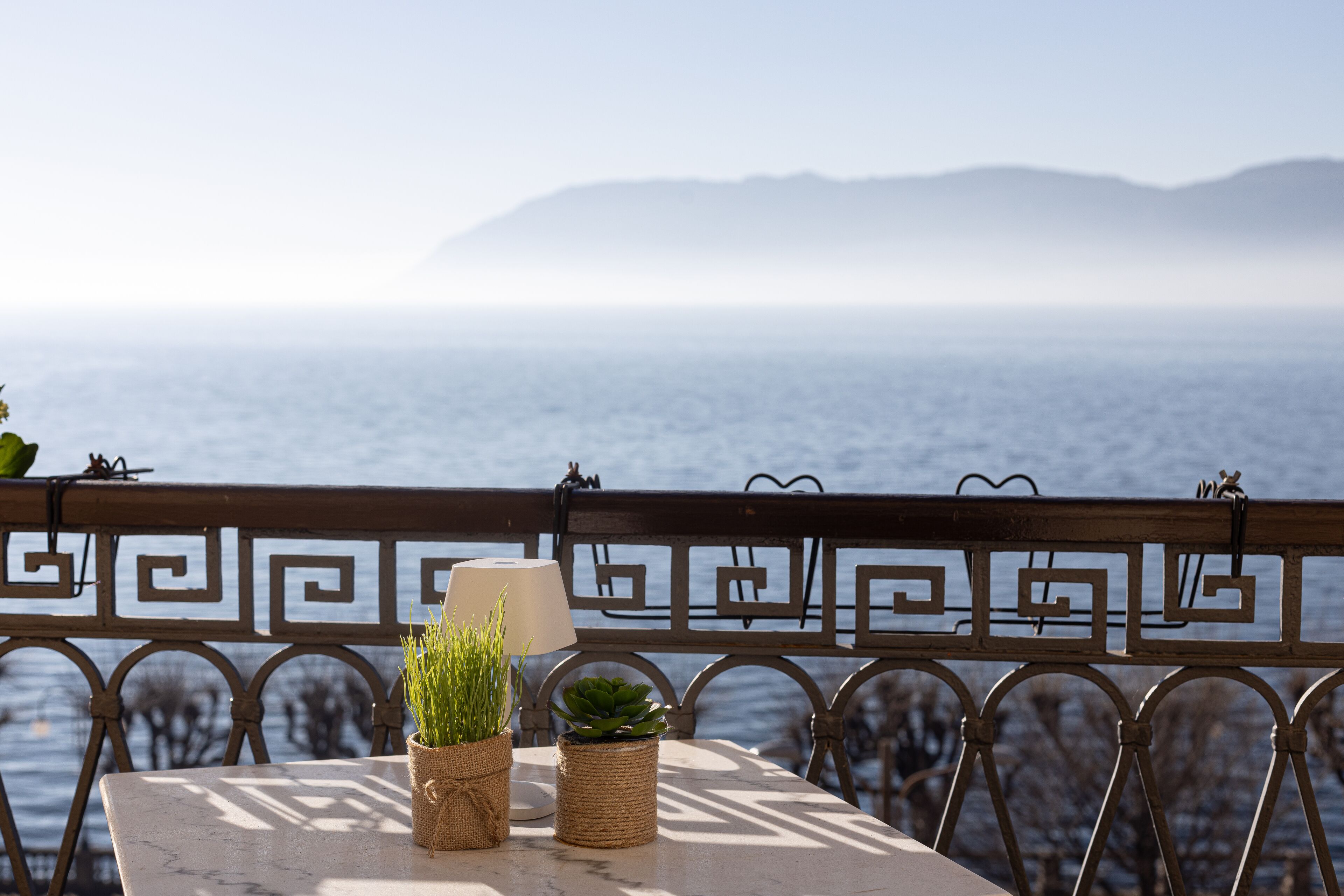 A table with a lamp and two potted plants, overlooking a body of water with mountains in the distance.