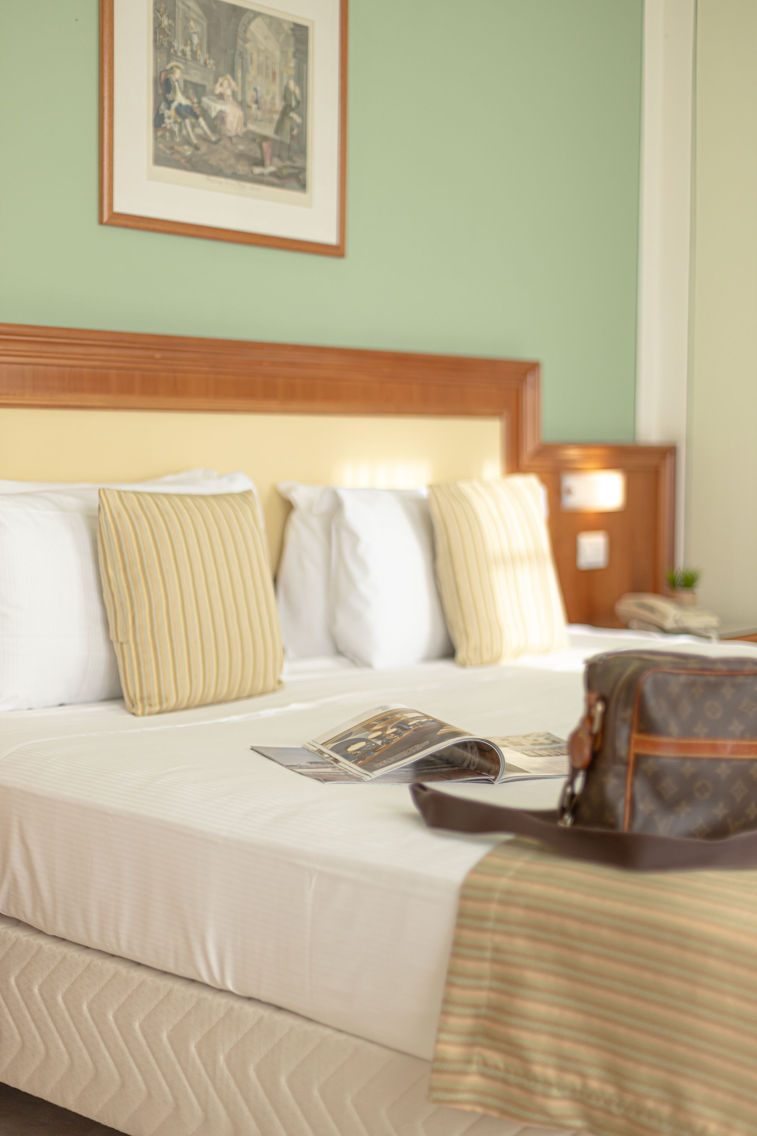 A neatly made bed with white bedding and striped pillows, a framed picture on the wall, and a brown bag on the bed.