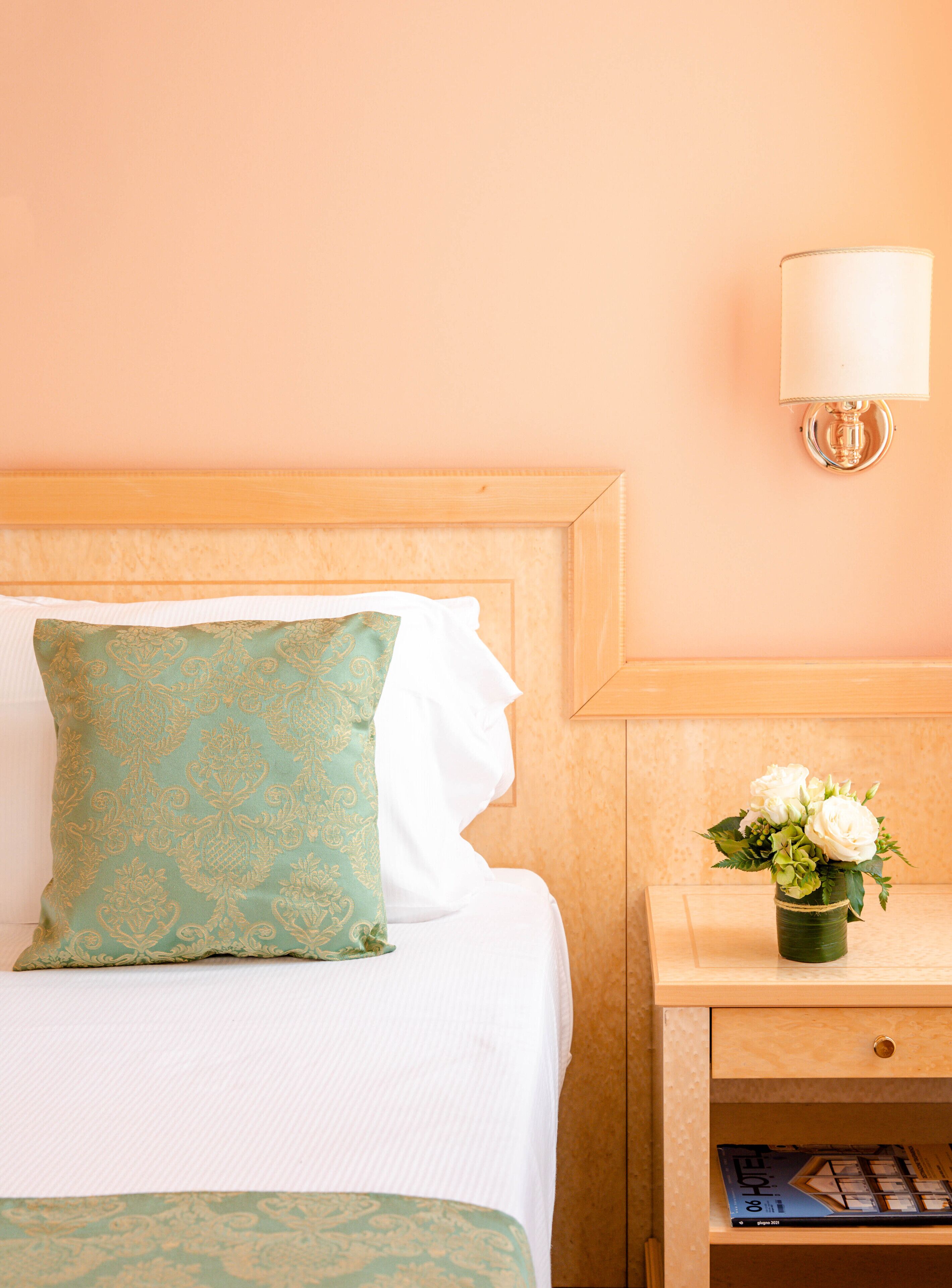 A neatly made bed with a patterned pillow and a white pillow, a bedside table with a lamp, and a small vase with flowers.