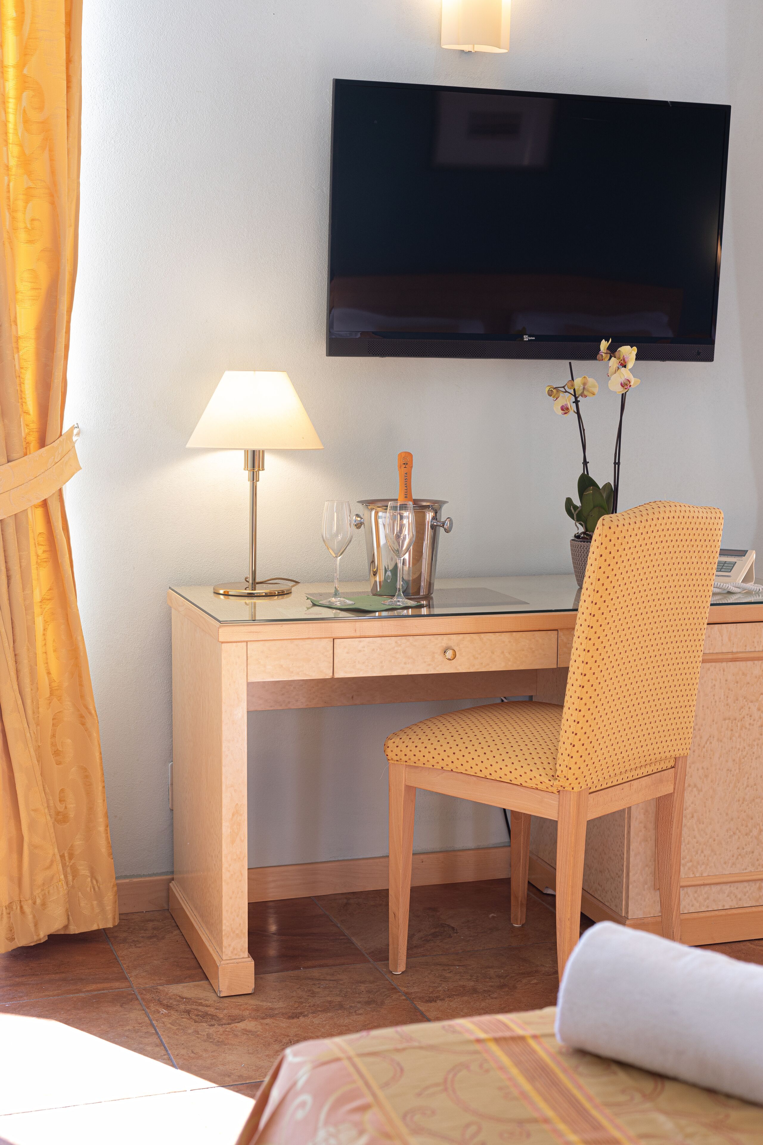 Quarto de hotel com secretária de madeira, televisão na parede, luminária, um vaso com flores e cama com colcha estampada.