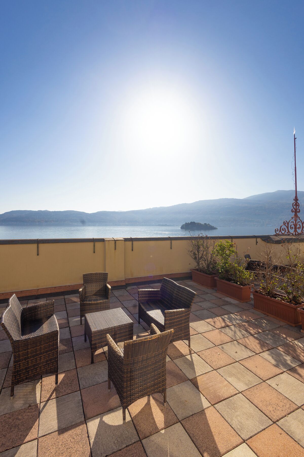 Una terraza con mobiliario de mimbre y plantas en macetas, con vistas a un cuerpo de agua y colinas distantes.