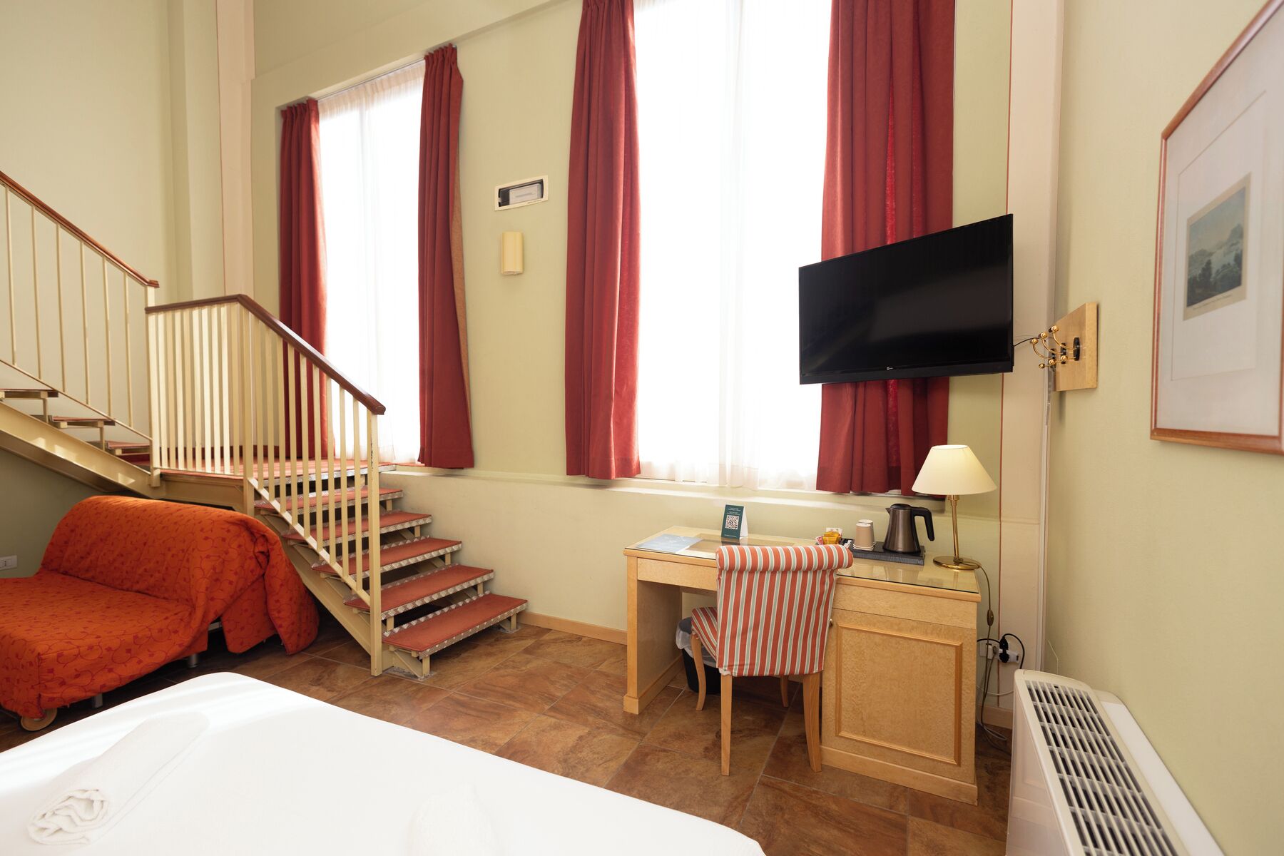 Une chambre d’hôtel comprenant un lit, un bureau, une chaise, une télévision, une fenêtre avec des rideaux, un escalier et un radiateur.