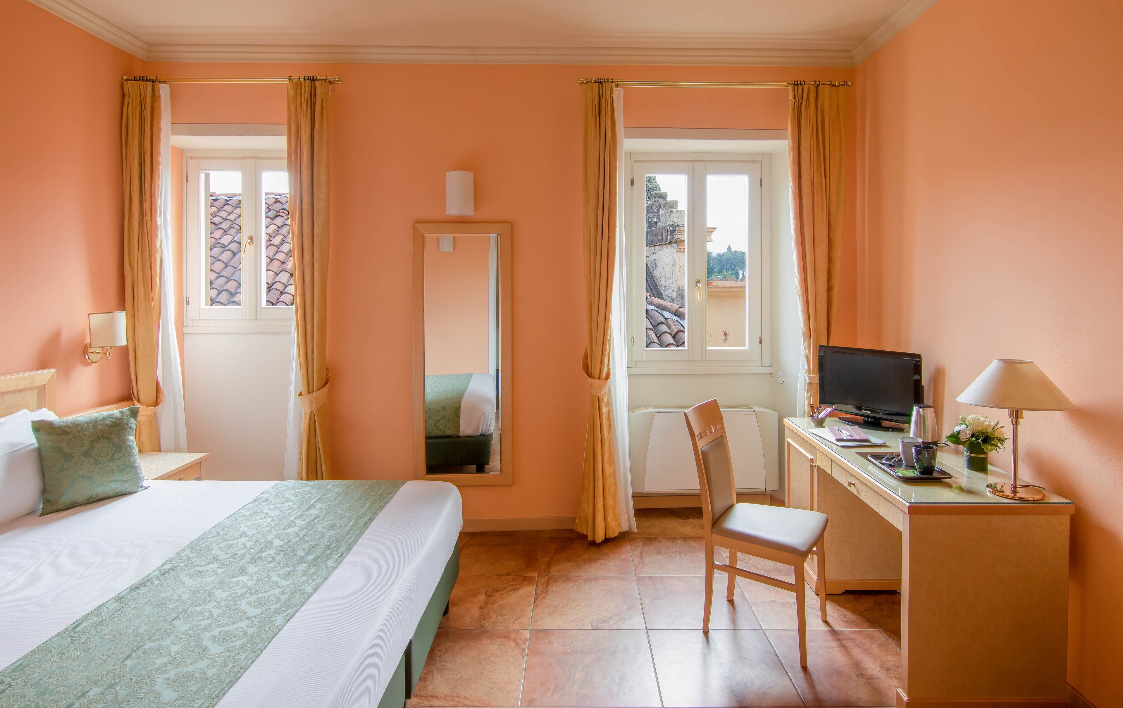 A hotel room with a bed, a desk with a computer, a chair, and a window with curtains.