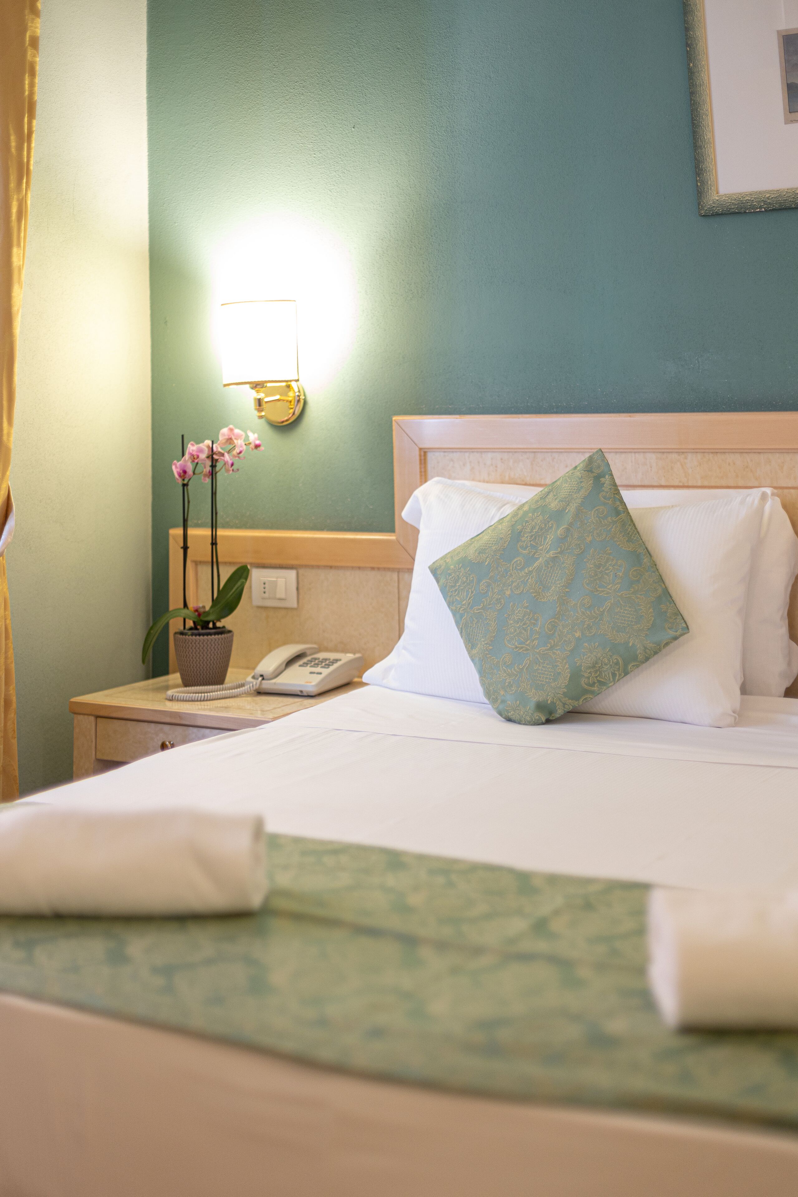 A neatly made bed with a green patterned pillow, a bedside phone, and a potted orchid on a nightstand.
