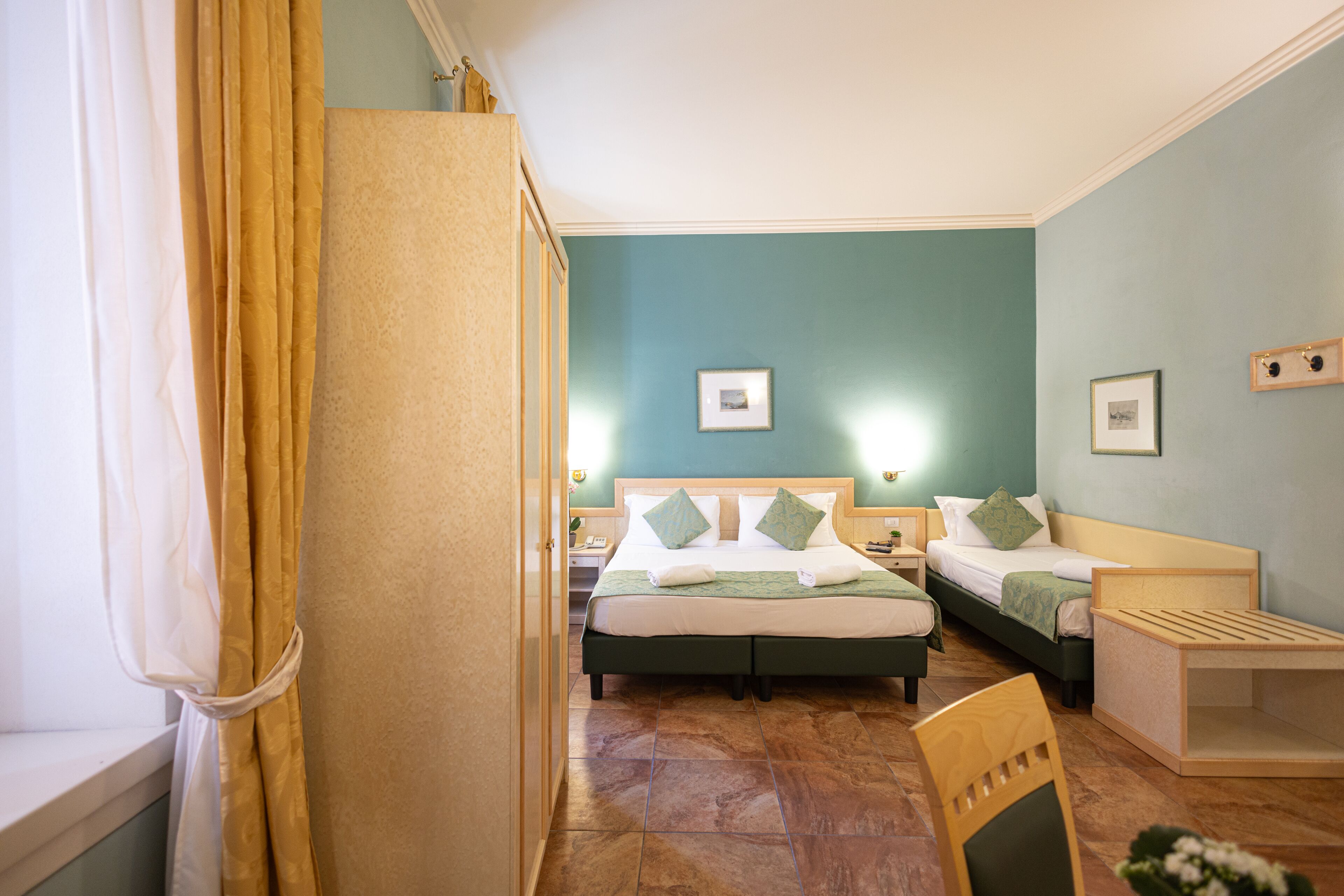 A hotel room with two beds, a wooden chair, a small table, and a window with curtains.