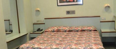 A hotel room with a bed, a nightstand, a bedside table, a mirror, and a framed picture on the wall.