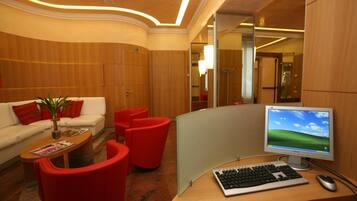 A modern hotel lobby with a curved wooden reception desk, a computer setup, red chairs, a white sofa with red cushions, and a small plant on a coffee table.