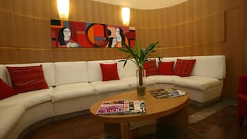A modern lounge area with a curved white sofa adorned with red pillows, a wooden coffee table with magazines, and a large abstract painting on the wall.