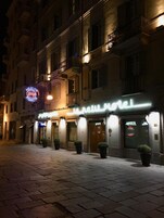A hotel with a neon sign, potted plants, and a brick-paved street at night.