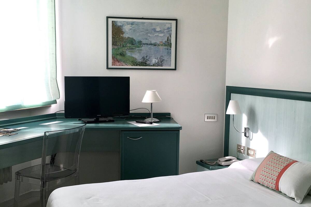 A hotel room with a bed, desk, chair, and a painting on the wall.