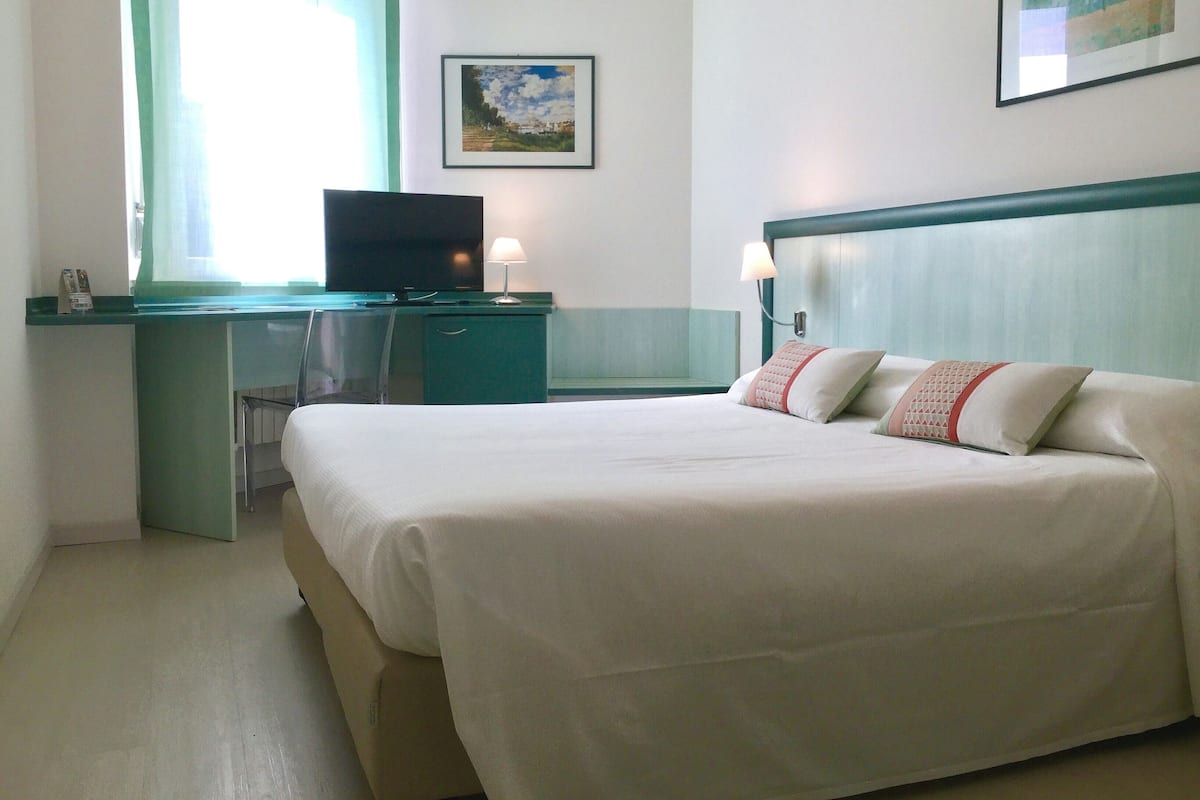 A hotel room with a bed, a desk, a television, and a painting on the wall.