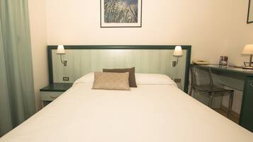 A neatly made bed with two pillows, a bedside table with a lamp, a desk with a chair, and a framed artwork on the wall.