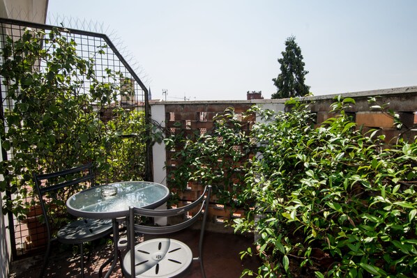 Un patio con una mesa redonda de vidrio y sillas metálicas, rodeado de abundante follaje verde y un muro de ladrillo.