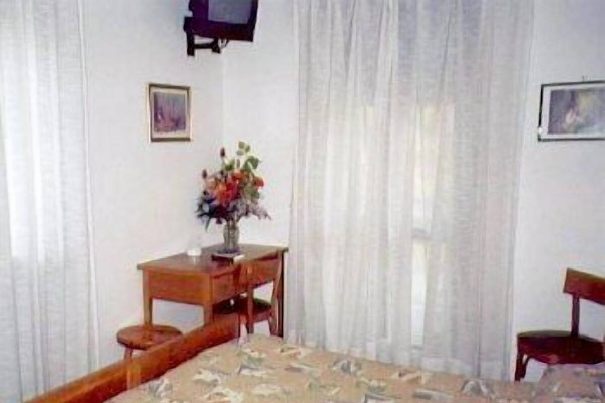 A bedroom with a bed, a wooden desk, a chair, a small table, and a vase with flowers.