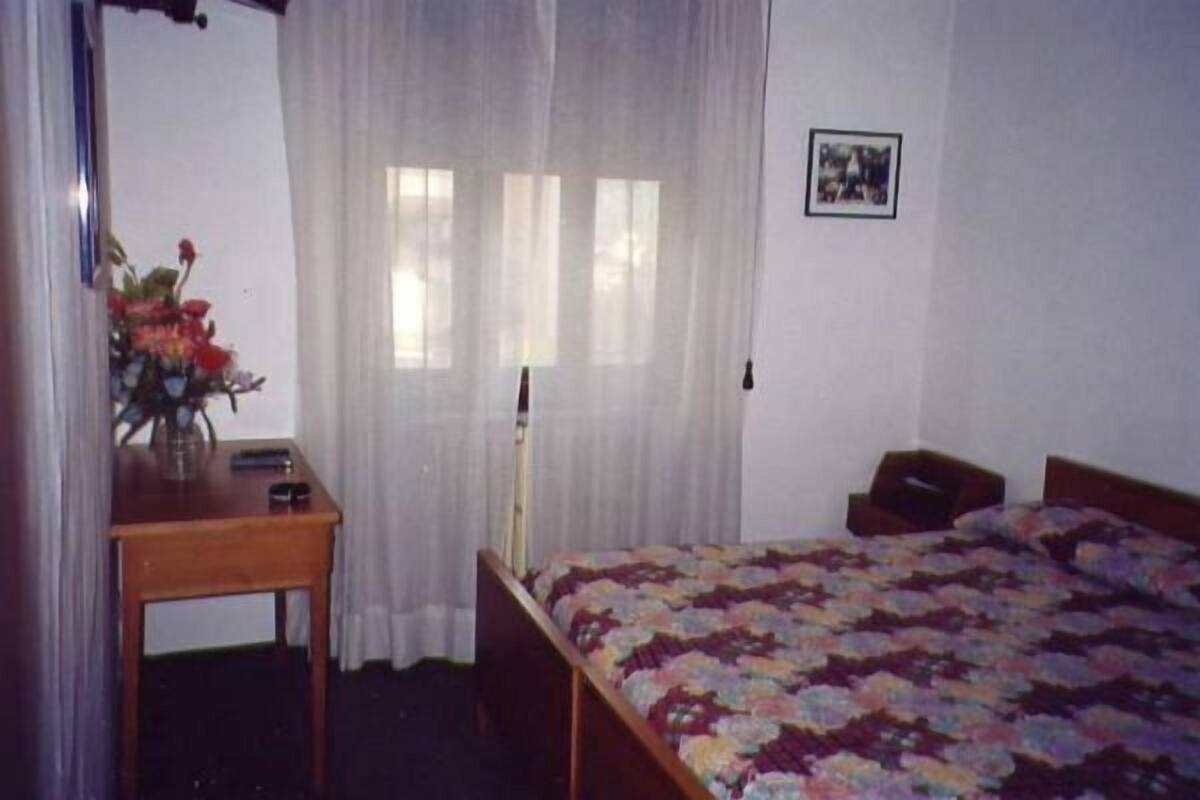 A bedroom with a bed, a wooden nightstand, a desk with a lamp, and a window with sheer curtains.