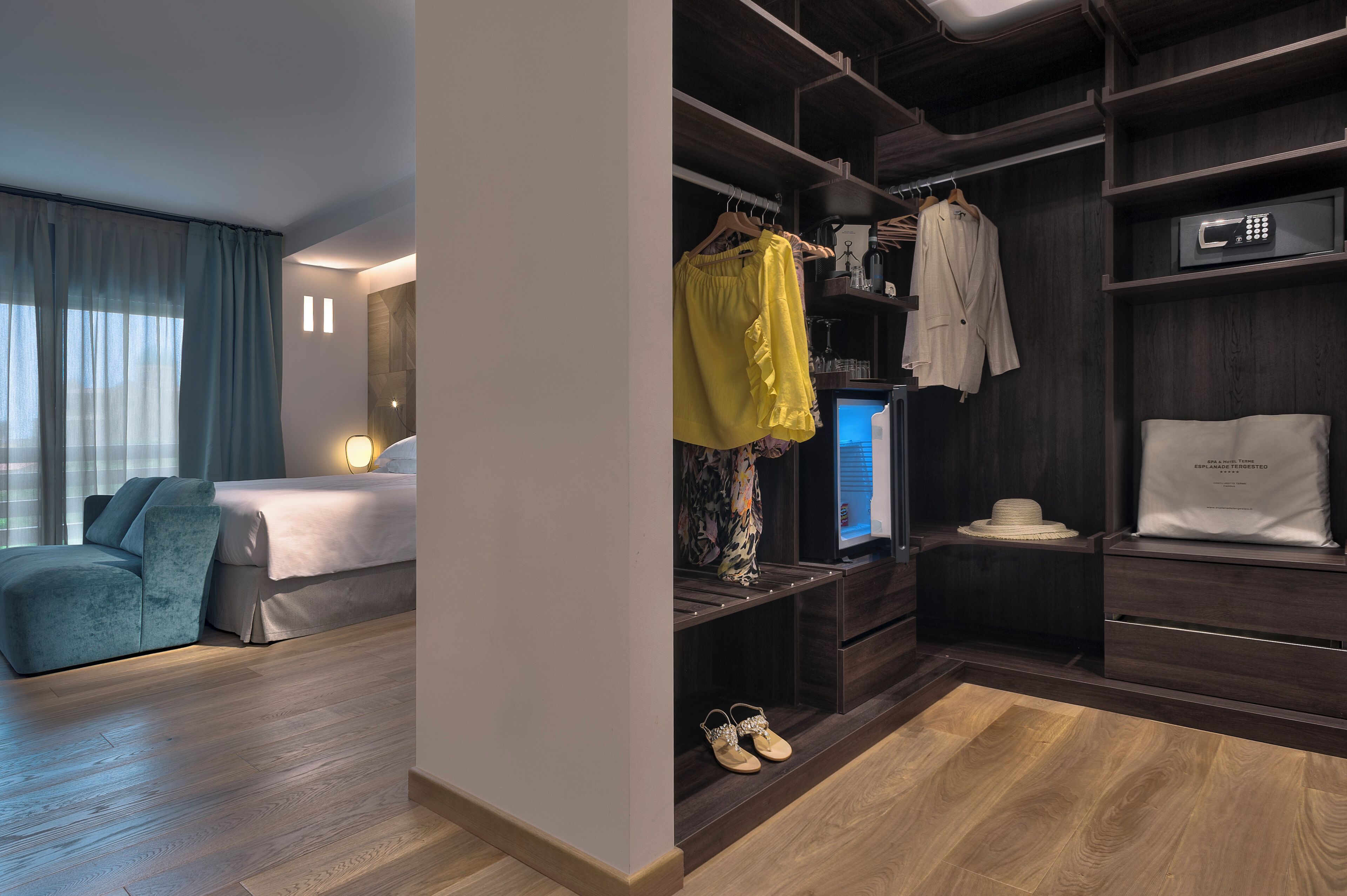 A hotel room with a bed, a seating area, a vanity with a mirror, and a wardrobe with hanging clothes and a suitcase.