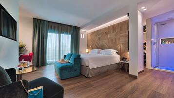Una habitación de hotel moderna con una cama grande, un área de descanso con sofá y sillón, una mesita con lámpara y un espacio para la ducha.