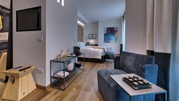 A modern hotel room with a sofa, a small table with a remote control, a glass coffee table, and a bed with a nightstand.