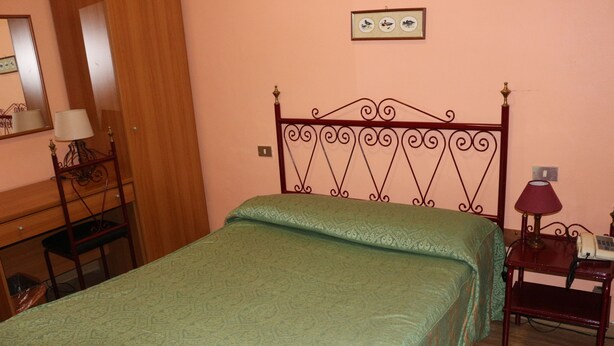 A bedroom with a green patterned bedspread, a wooden nightstand with a lamp, and a wall-mounted picture. - Hotel Italia (Turin)